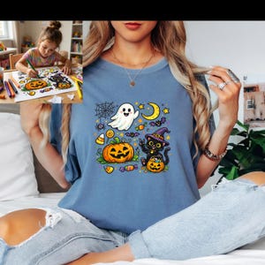 May include: A blue t-shirt featuring a colorful Halloween graphic print of a smiling ghost, a black cat wearing a purple witch hat, a jack-o'-lantern, a spider web, bats, stars, a crescent moon, and various candies scattered across the center.