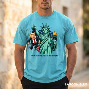 May include: Lagoon blue t-shirt featuring a graphic of the Statue of Liberty holding a cartoon figure by the scruff of the neck. The graphic includes the text "USA THIS IS NOT A KINGDOM".
