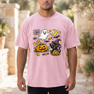 May include: A light pink short-sleeve t-shirt featuring a colorful Halloween graphic. The design includes a white ghost, a jack-o'-lantern, a black cat wearing a purple witch hat, a spider web, a yellow crescent moon, and various candies.