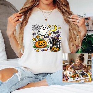May include: A cream-colored t-shirt featuring a colorful Halloween graphic print. The design includes a smiling ghost, a jack-o'-lantern, a black cat wearing a witch hat, a spider web, a crescent moon, stars, bats, and various candies.