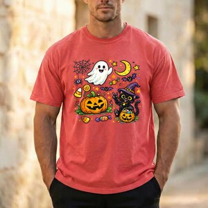 May include: A red short-sleeve t-shirt featuring a colorful Halloween graphic print. The design includes a white ghost, a large orange jack-o'-lantern, a black cat wearing a purple witch hat, a yellow crescent moon, stars, and candy.