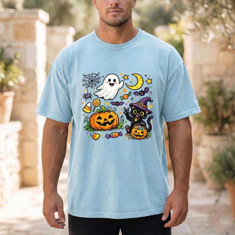 May include: A light blue t-shirt featuring a colorful Halloween graphic. The design includes a white ghost, a jack-o'-lantern, a black cat wearing a purple witch hat, a spider web, a crescent moon, stars, bats, and various pieces of candy.