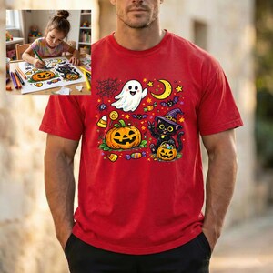 May include: A red t-shirt featuring a colorful Halloween graphic print of a white ghost, a black cat wearing a purple witch hat, a jack-o'-lantern, a yellow crescent moon, stars, spiders, and various candies scattered across the front.