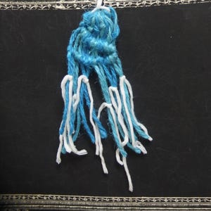 May include: A handmade bookmark featuring a blue and white yarn design. The top portion is a tightly woven blue knot, with long strands of blue and white yarn hanging below. The bookmark is set against a dark background.