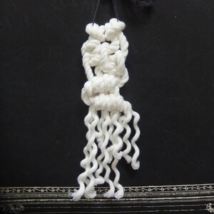 May include: A handmade macrame wall hanging featuring a knotted design with cascading, wavy white fringe. The top has a black cord for hanging. The background is a dark surface with decorative border.