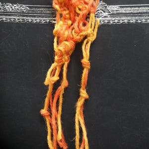 May include: A handmade macrame piece in shades of orange and yellow. The knotted design features a central cluster of intertwined cords, with several strands extending downwards. The piece is crafted from thick, textured yarn.