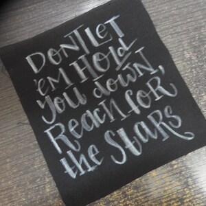 Op de afbeelding: Een vierkante zwarte stoffen patch met witte handgeschreven kalligrafie met de tekst: Don't let 'em hold you down, reach for the stars. De letters hebben een penseelstreektextuur en de stof heeft zichtbare gerafelde randen aan de zijkanten.