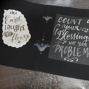 May include: A black fabric piece featuring two white graphic designs. The left design is a white cutout with the text "the Earth laughs in flowers". The right design is a black square with white text reading "COUNT your Blessings NOT YOUR PROBLEMS".