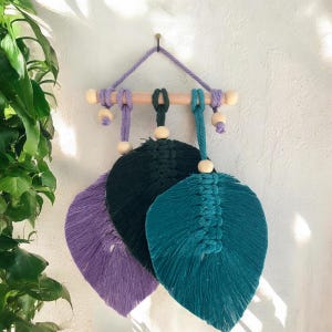 May include: A wall hanging featuring three macrame leaves in purple, dark green, and teal. The leaves are suspended from a light-colored wooden dowel by matching colored cords, accented with natural wooden beads and a purple hanging cord.