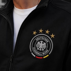 Può includere: Una giacca sportiva nera con zip che presenta il logo della Deutscher Fussball-Bund ricamato sul petto. L'emblema mostra un'aquila centrale, quattro stelle dorate sopra e una fascia curva con i colori nero, rosso e giallo alla base.