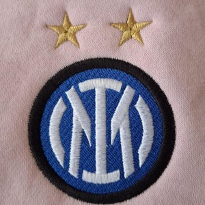 May include: A light pink fabric featuring an embroidered circular Inter Milan football club logo in blue, white, and black. Above the circular emblem, two gold-colored embroidered stars are positioned side by side on the pink textile surface.