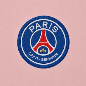 May include: A circular embroidered patch featuring the Paris Saint-Germain logo on a light pink fabric background. The emblem displays a red Eiffel Tower silhouette, a gold fleur-de-lis, and white text on a blue field with a white border.