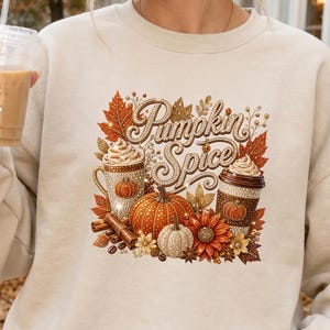 May include: A beige crewneck sweatshirt featuring a graphic print of two coffee cups, pumpkins, cinnamon sticks, and autumn leaves. The text "Pumpkin Spice" is written in a decorative script font. The design includes sparkling accents throughout.