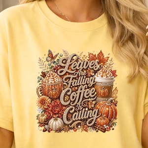 May include: A pale yellow t-shirt featuring a central graphic with the text "Leaves Are Falling Coffee Is Calling" in a glittery font. The design includes two coffee cups, orange pumpkins, autumn leaves, a butterfly, and cinnamon sticks.