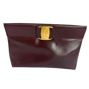 May include: A dark burgundy leather clutch bag featuring a horizontal grosgrain ribbon band across the top. Centered on the ribbon is a polished gold-tone metal Vara bow plaque engraved with the Salvatore Ferragamo brand name.