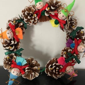 May include: A handmade Christmas wreath crafted from pinecones, adorned with small elf figures, colorful felt accents, and red, green, and gold embellishments. The wreath is designed in a circular shape, perfect for holiday decor.