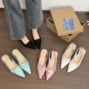 Puede incluir: Se muestran varios pares de zapatos de tacón con punta y correa trasera, con varios diseños. Los zapatos incluyen diseños florales negros, florales azul claro, florales rosas y encaje blanco. También se ve una caja marrón con una tarjeta azul.