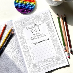 May include: An open coloring book with the title "Focus Flow Vol. 1" and the words "Dopamine boost" is next to a rainbow-colored pop-it toy, a cup of coffee, and several colored pencils. The book has floral designs.