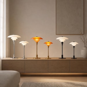 Może przedstawiać: Kolekcja nowoczesnych lamp stołowych z warstwowymi kloszami. Lampy mają metalowe, mosiężne i czarne podstawy. Klosze są białe i pomarańczowe, tworząc ciepłe światło. Lampy są ustawione na drewnianej szafce.