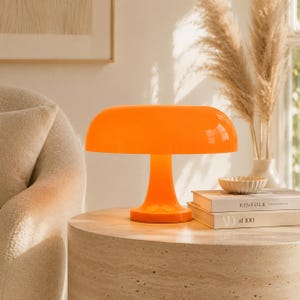 May include: An orange mushroom-shaped table lamp with a rounded shade and a pedestal base. The lamp is on a round, light-colored table next to books and a decorative bowl. The lamp's bright color contrasts with the neutral tones of the room.