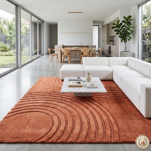 May include: A modern living room featuring a large, rust-colored rug with a concentric circle design. The rug sits beneath a white sectional sofa and a marble coffee table. The room has large windows and a minimalist aesthetic.