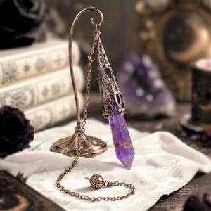 Può includere: Un pendolo di cristallo viola con un involucro e una catena in metallo decorativi, esposto su un supporto in bronzo. Il cristallo è di un viola intenso con striature più chiare e il metallo ha un design a filigrana ornato. Il pendolo è appoggiato su un panno bianco.