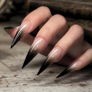 Könnte beinhalten: Eine Hand mit langen, spitzen Fingernägeln. Die Nägel haben ein Farbverlauf-Design, das von einem nackten Grundton zu einem tiefen Schwarz an den Spitzen übergeht. Die Nägel sind glänzend und gepflegt, was einen dramatischen und stilvollen Look erzeugt.
