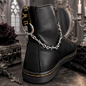 Könnte beinhalten: Schwarzer Leder-Ankle-Boot mit gelb genähter Sohle. Eine silberne Kette mit Stacheldraht-Design und einem Totenkopf-Anhänger ist an der Stiefel befestigt. Der Stiefel hat eine gelb-schwarze Zuglasche.