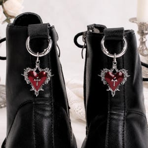 Puede incluir: Primer plano de dos botines de cuero negro. Cada bota presenta un anillo plateado en la parte superior, con un dije en forma de corazón. Los dijes tienen un centro rojo con una cruz y están rodeados por un borde plateado decorativo.
