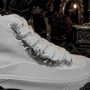 Op de afbeelding: Een witte canvas high-top sneaker met een zilveren vleermuisvormige schoenbedel. De bedel heeft gedetailleerde vleugel- en lichaamskenmerken. De veters zijn wit en de vetergaten zijn zilver.