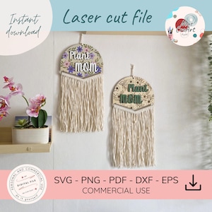 May include: Two semi-circular wooden wall hangings featuring the text Plant mom in raised lettering. The left piece has a floral pattern with purple flowers, while the right has a black line-art floral design. Both have cream macrame fringe.
