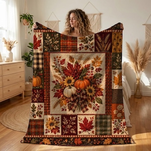 May include: A rectangular throw blanket featuring a central panel of autumn harvest imagery including pumpkins, sunflowers, and fall leaves, surrounded by a patchwork border of plaid patterns, foliage, and berries in shades of orange, red, and brown.