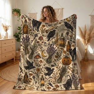 May include: A cream-colored throw blanket featuring a repeating pattern of black cats, crows, pumpkins, glass apothecary bottles, lavender bundles, mushrooms, keys, spiderwebs, and crescent moons in shades of brown, gold, and purple.