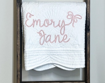 以下が含まれることがあります： ピンク色の刺繍で「Emory Jane」とリボン模様が施された白いベビーブランケット。ブランケットは木製の梯子に折りたたまれており、キルトの質感と装飾的なステッチが特徴です。