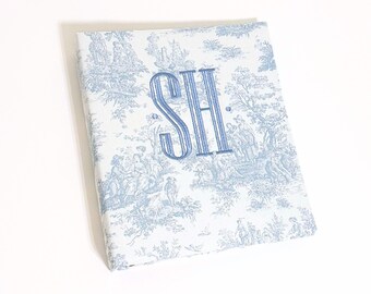 May include: A white fabric binder with a blue toile pattern and the monogram "SH" embroidered in blue.