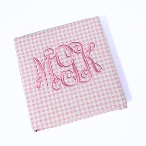 May include: A pink and white gingham pattern notebook with a pink embroidered monogram of the letters MGK. The notebook is closed and angled, with a shadow visible on a white background.