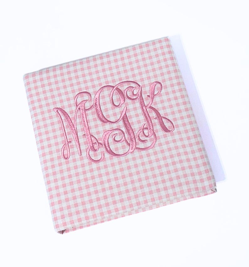 May include: A pink and white gingham pattern notebook with a pink embroidered monogram of the letters MGK. The notebook is closed and angled, with a shadow visible on a white background.