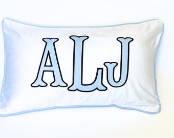 以下が含まれることがあります： 白の長方形の枕で、水色の縁取りがあります。「ALJ」の文字が水色で黒い縁取りで描かれています。文字は枕の中央に配置されています。