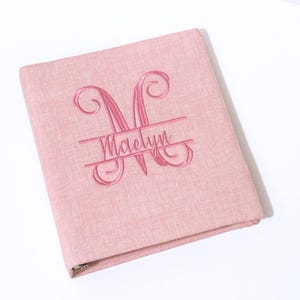 May include: A pink binder with an embroidered monogram and the name "Maelyn". The binder has a textured fabric cover and a metal ring binder mechanism. The monogram is a stylized "M".