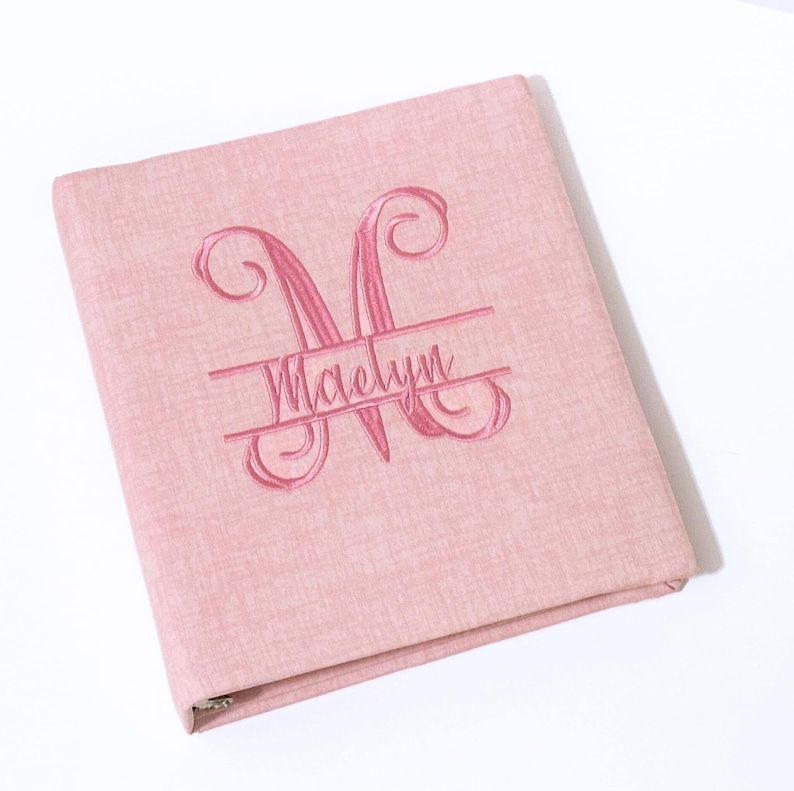 May include: A pink binder with an embroidered monogram and the name "Maelyn". The binder has a textured fabric cover and a metal ring binder mechanism. The monogram is a stylized "M".