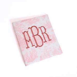 May include: A square, light pink and white toile notebook with a red embroidered monogram. The notebook features a classic toile pattern and a three-letter monogram in a bold, elegant font. The cover is slightly angled.