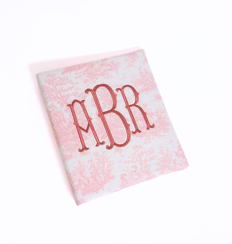 May include: A square, light pink and white toile notebook with a red embroidered monogram. The notebook features a classic toile pattern and a three-letter monogram in a bold, elegant font. The cover is slightly angled.