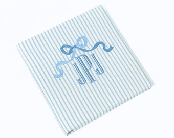 May include: A folded, striped blanket with blue and white vertical stripes. The blanket features a light blue embroidered bow and the initials "JPJ" in a matching light blue thread.