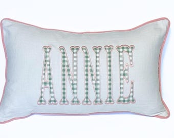以下が含まれることがあります： 緑と白のギンガムチェックの生地に「ANNIE」という名前が刺繍された長方形の枕。 枕は薄い青色の背景にピンク色のトリムが付いています。