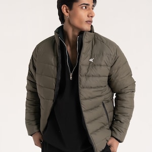 Puede incluir: Una chaqueta acolchada de color verde oliva con costuras horizontales, cuello alto y cierre de cremallera frontal. La chaqueta presenta un pequeño logotipo blanco en el pecho. Debajo, una prenda negra con cremallera y pantalones negros con el texto BLACKHAWK DEFY THE LIMITS.