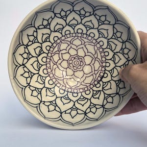 May include: A round ceramic bowl with a glossy off-white finish, featuring a detailed black ink mandala design that radiates from the centre. The intricate pattern consists of symmetrical petal-like shapes and concentric circles covering the interior.