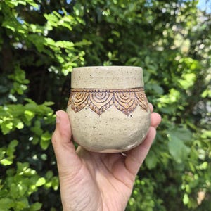 May include: A speckled beige ceramic cup featuring a hand-carved decorative band of repeating scalloped arch patterns around the upper section. The pottery has a rounded, bulbous shape with a smooth finish and dark brown detailing in the carvings.