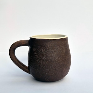 May include: A dark brown ceramic mug with a rounded body and a curved handle. The exterior surface features an intricate, embossed mandala-style floral pattern. The interior of the mug is finished in a smooth, contrasting off-white glaze.