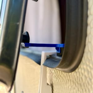 May include: A blue plastic window stay latch mechanism installed on a caravan window frame. The device features a horizontal arm that holds the dark-tinted window pane open, secured against a white plastic mounting bracket on the textured wall.