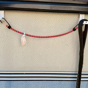 May include: Two white plastic wall-mounted hooks attached to a textured surface, connected by a red and black bungee cord with metal hooks at each end. A black nylon strap hangs from the right hook, and a small white tag is attached to the cord.